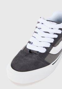 Baskets en suede grises avec lacets blancs et accents latéraux, présentant un dessus texturé, une pointe arrondie et une semelle en caoutchouc blanche.
