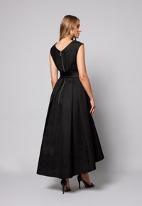 Czarna sukienka bez rękawów z plecami w kształcie litery V, długą plisowaną spódnicą i wiązaniem w talii. Posiada zamek błyskawiczny z tyłu i teksturowany materiał. W zestawieniu z wysokimi obcasami.