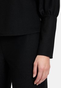 Schwarzes, strukturiertes Oberteil mit lässigem Schnitt und Puffärmeln, kombiniert mit passenden schwarzen Hosen. Der Stoff hat einen dezenten Glanz.