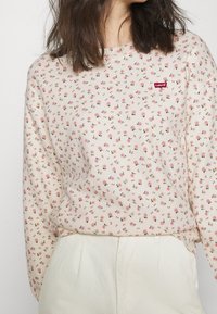 Maglietta a maniche lunghe color crema con stampa floreale, caratterizzata da piccoli fiori rosa e verdi, e il logo rosso di Levi's sul petto. Tessuto dalla superficie liscia.