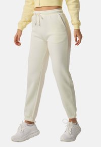 Žena v krémových teplácích s tkaničkou v pase a bílými teniskami stojící na bílém pozadí.