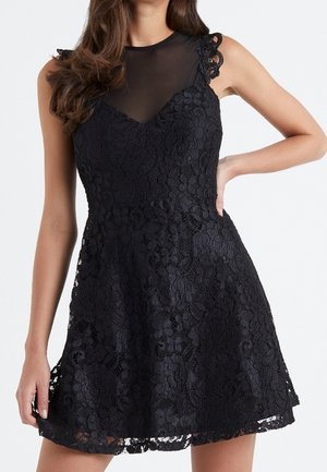 Robe noire sans manches en dentelle avec motif floral, empiècement en maille transparente, corsage ajusté et jupe évasée, portée par une femme debout.