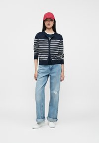 Νεαρή γυναίκα που φοράει κόκκινο καπέλο, ναυτικό ριγέ ζακέτα πάνω από σκοτεινό T-shirt, ανοιχτό μπλε σκισμένα τζιν και λευκά αθλητικά παπούτσια, που στέκεται σε λευκό φόντο.