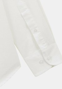 Chemise blanche à manches longues avec un col pointu, dotée d'un poignet à bouton et d'une texture lisse. Une transparence visible dans le tissu et des coutures minimales.