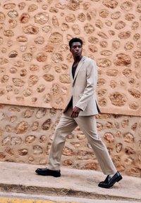 Homme en costume beige et chaussures noires marchant sur un pavé en pierre devant un mur en pierre texturé.