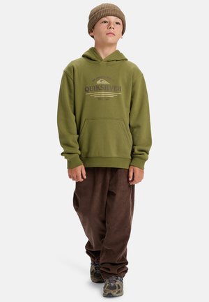 Haut à capuche vert olive avec poche avant et logo imprimé, associé à un pantalon en velours côtelé marron et des baskets à motifs. Bonnet en tricot chaud sur la tête.