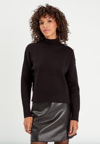 Pull en laine noir avec un col haut et un ourlet côtel�é, associé à une jupe en cuir noir. Présente un accent matériel subtil sur l'épaule.
