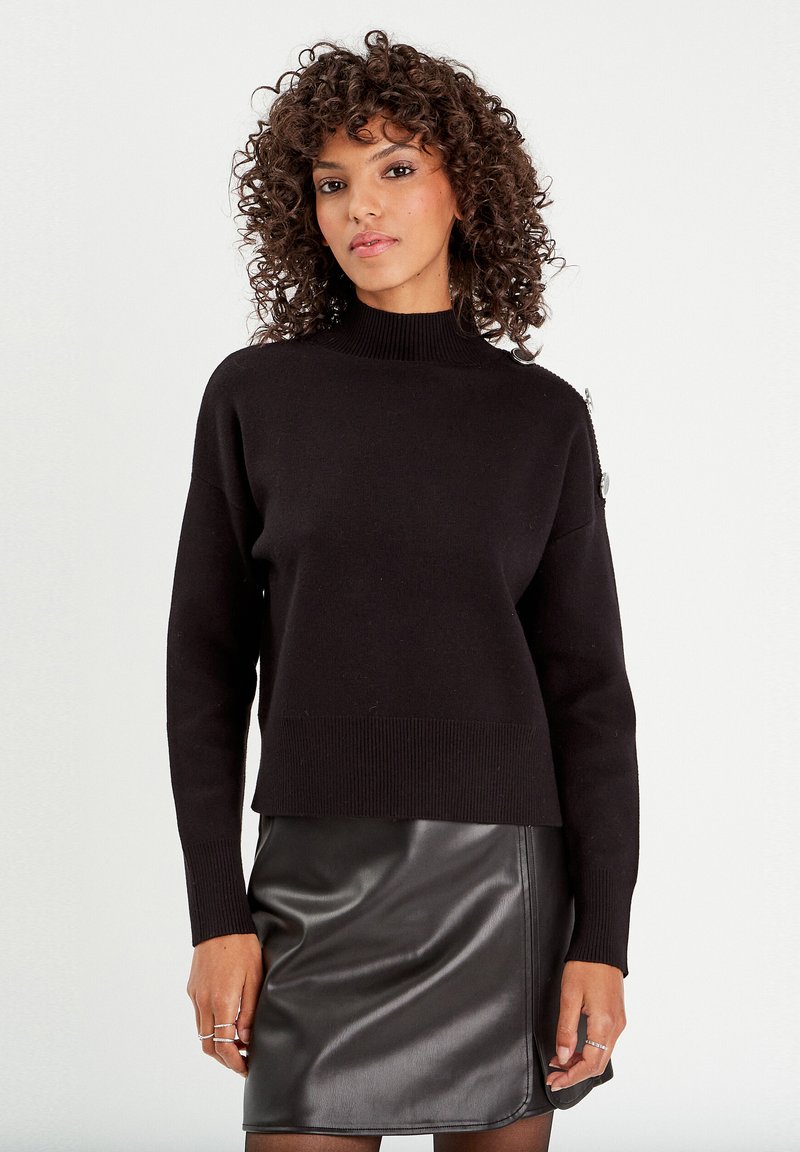 Pull en laine noir avec un col haut et un ourlet côtelé, associé à une jupe en cuir noir. Présente un accent matériel subtil sur l'épaule.