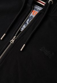 Schwarzer Kapuzenpullover mit Reißverschluss und weicher Textur, ausgestattet mit silberner Reißverschluss-Hardware, gesticktem Markenlogo und kontrastierendem Innenlabel-Design.