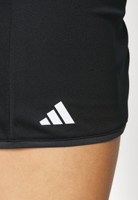 Juodi sportiniai šortai iš kvėpuojančios medžiagos, su baltu Adidas logotipu ir lygiu kraštu.