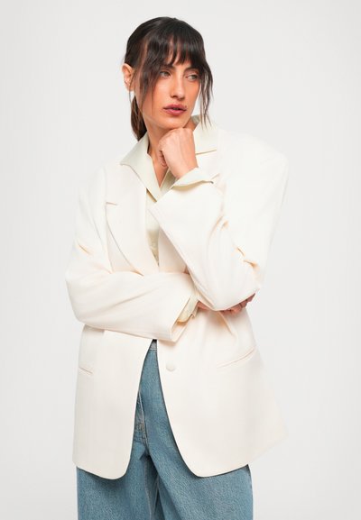 Vrouw met donker haar in een lage paardenstaart draagt een oversized crèmekleurige blazer over een licht shirt en blauwe jeans, poseert met haar hand bij haar kin tegen een witte achtergrond.
