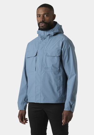 Mężczyzna ubrany w jasnoniebieską kurtkę z kapturem i kieszeniami z patkami, stojący na jednolitym jasnym tle, patrzący przed siebie.