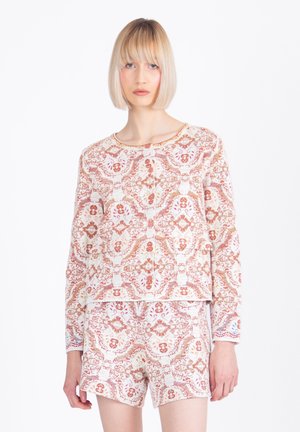 Ženska s kratkimi blond lasmi, oblečena v ujemajočo dolgoročno srajco z gumbi in kratke hlače z zapletenimi rdečimi in belimi cvetličnimi vzorci.