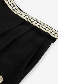 Schwarzes Kleidungsstück mit einem gefältelten Design, verziert mit einem dekorativen silbernen und weißen Besatz am Ausschnitt. Weiche Textur, lässiger Look.