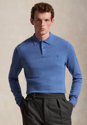 Jeune homme aux cheveux bouclés portant un polo bleu à manches longues et un pantalon plissé taille haute gris foncé, les mains dans les poches.