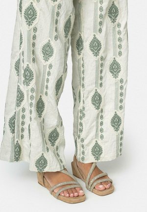 Vista desde las piernas de una persona que lleva pantalones anchos crema con un estampado floral verde y sandalias beige de punta abierta con tres tiras delgadas.