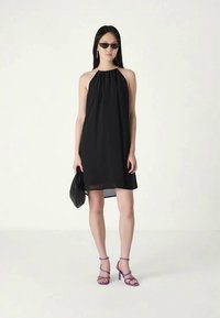 Mulher vestindo um vestido halter preto sem mangas, sapatos de salto roxos com tiras, óculos de sol pretos, segurando uma bolsa de mão preta, de pé contra um fundo simples.