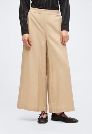 Femme portant un pantalon beige à jambes larges, une chemise noire à fines rayures et des ballerines noires à lacets, debout devant un fond uni.
