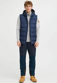 Navy puffer vest with a hood, quilted design, paired with a light gray long-sleeve top and dark cargo pants. Tan lace-up boots complete the look.