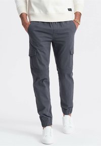 Homme portant un pantalon jogger cargo gris foncé et des baskets blanches, les mains dans les poches, sur fond uni.