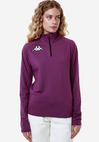 Maglia atletica a maniche lunghe, con zip e collo alto, di colore viola, con una finitura testurizzata; presenta un logo bianco sul petto sinistro e maniche aderenti.