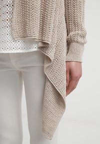 Světlý béžový pletený cardigan s asymetrickým lemem a žebrovanými manžetami, vrstvený přes bílý vzorovaný top, doplněný světlými kalhotami.