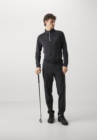 Black long-sleeve zip-up shirt paired with black pants and a belt. The individual holds a golf club, wearing black athletic shoes.