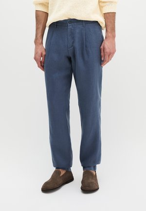 Mężczyzna noszący niebieskie plisowane spodnie, beżowy sweter i brązowe zamszowe buty wsuwane, stojący na jasnym jednolitym tle.