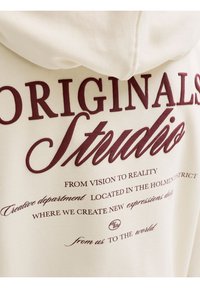 Φούτερ με κουκούλα με μπορντό κείμενο "ORIGINALS Studio" και επιπλέον φράσεις σχετικά με το δημιουργικό τμήμα και νέες εκφράσεις σε μπεζ ύφασμα.