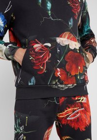 Svart blommig sweatshirt och byxset med livfulla röda, blåa och vita blommönster. Har en framficka och ribbad mudd. Blandning av bomullsmaterial.