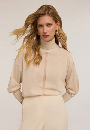 Maglione beige a collo alto con dettaglio in pannello trasparente, vestibilità ampia, polsini a coste e texture morbida, abbinato a pantaloni color crema.