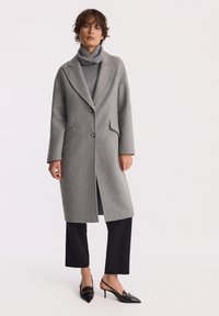 Kobieta ubrana w szary długi płaszcz na szary golf, czarne spódnico-spodnie i czarne czółenka na wysokim obcasie, stoi na białym tle.
