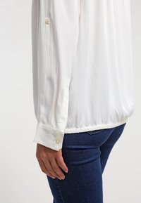 White, long-sleeve blouse made of lightweight fabric, featuring a button detail on the cuffs and an elastic hem. Worn with dark blue jeans.