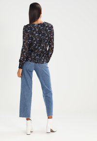 Donna con lunghi capelli scuri, che indossa una blusa nera a maniche lunghe con fiori, jeans blu a vita alta e stivaletti bianchi, in piedi e di spalle.