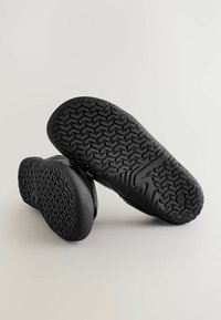Čierna obuv s textúrovanými gumovými podrážkami s cik-cak vzorom; hladký horný materiál s okrúhlou špičkou, ktorý má minimálne detaily.