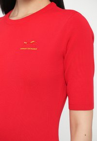 Κοντινό πλάνο κόκκινης κοντομάνικης πλεκτής μπλούζας με στρογγυλή λαιμόκοψη και μικρό κίτρινο κεντημένο λογότυπο Armani Exchange στο στήθος.