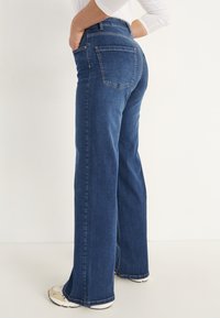 Kvinna klädd i högmidjade, raka mörkblå jeans med bakfickor, i kombination med vita sneakers och en vit långärmad tröja.