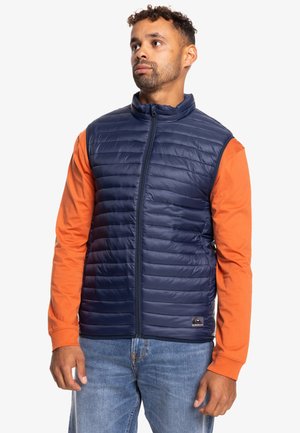 Marineblauwe gewatteerde vest met hoge kraag, voorrits en zijzakken, gedragen over een oranje overhemden met lange mouwen en gestyled met blauwe jeans.