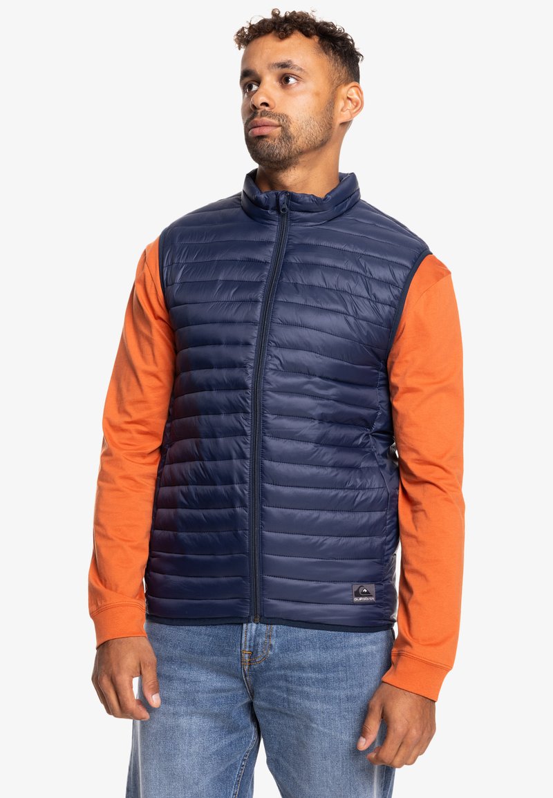Chaleco acolchado navy con cuello alto, cierre frontal y bolsillos laterales, usado sobre una camiseta de manga larga naranja y combinado con jeans azules.