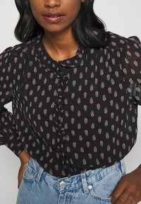 Sort bluse med blomsterprint i rød og hvid, med flæset krave og knaplukning, parret med lyseblå højtaljede jeans.