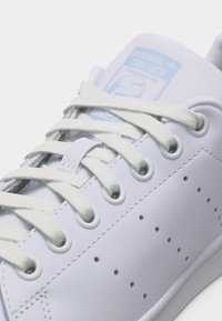 Κοντινό πλάνο λευκού αθλητικού παπουτσιού Adidas Stan Smith που δείχνει τα κορδόνια, τις τρύπες, τις τρύπες στις πλάγιες ρίγες και την ετικέτα στη γλώσσα με το λογότυπο σε ανοιχτό μπλε χρώμα.