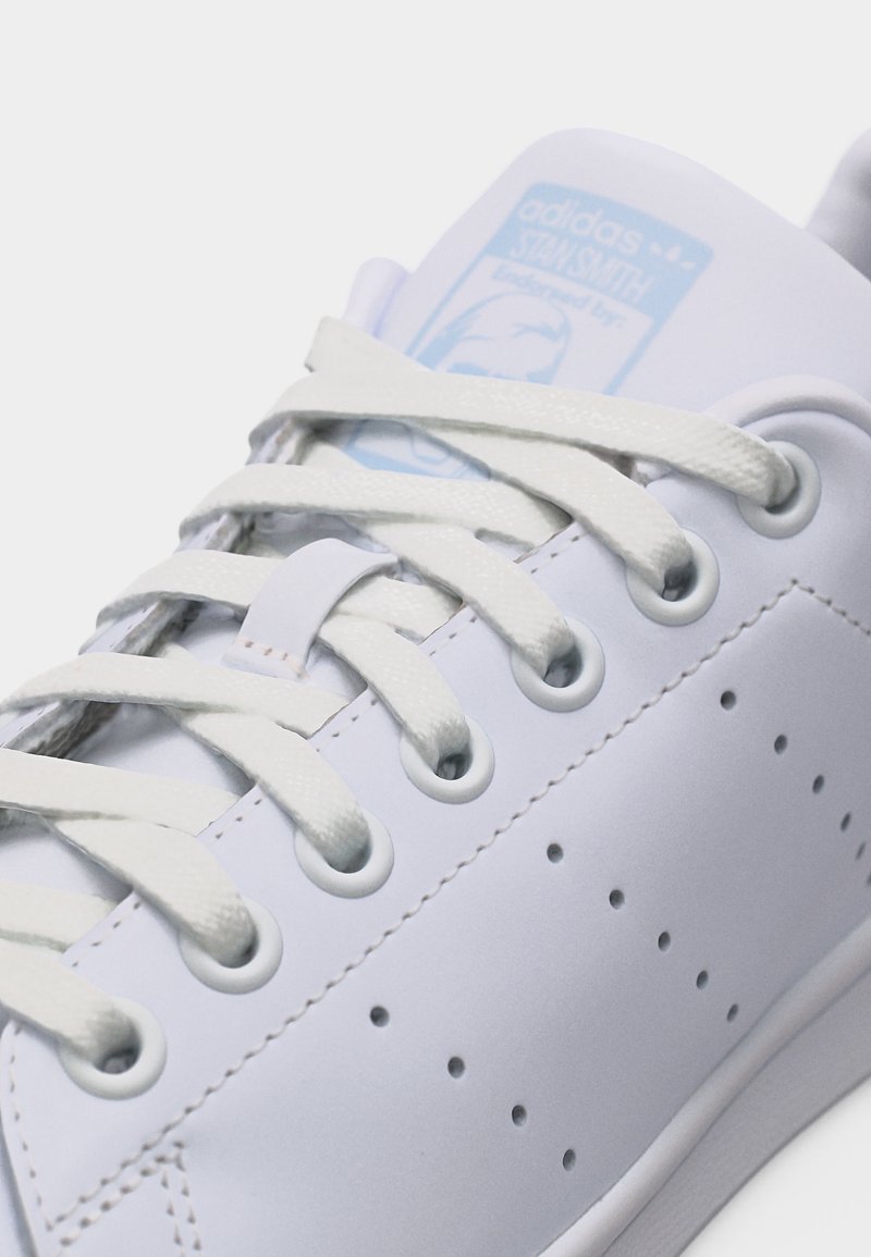 Nærbillede af hvid Adidas Stan Smith sneaker, der viser snørebånd, øjer, perforerede sidestriber og mærket tungemærke i lyseblå.