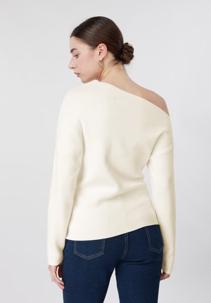 Femme avec les cheveux en chignon portant un pull blanc à épaules dénudées et un jean bleu foncé, vue de dos sur un fond clair uni.