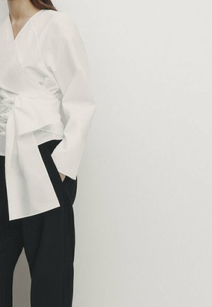 Femme portant un chemisier blanc cache-cœur avec de longues manches volumineuses et un pantalon noir à jambes larges, debout avec une main dans la poche.
