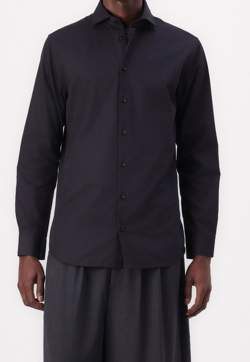 Homme portant une chemise noire à manches longues boutonnée et un pantalon ample gris foncé sur un fond uni.