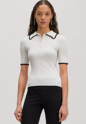 Frau mit langen lockigen Haaren trägt ein weißes kurzärmeliges, geripptes Oberteil mit schwarz eingefasstem Kragen und Manschetten, kombiniert mit schwarzen Hosen.