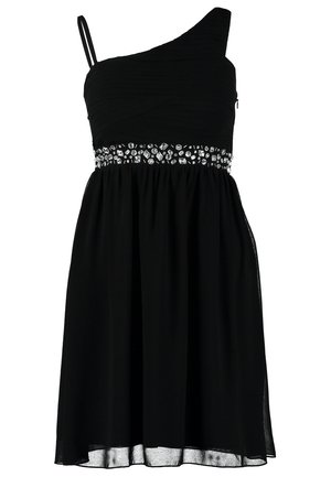 Femme portant une robe noire asymétrique avec une taille ornée de bijoux, tenant une pochette noire et portant des talons hauts noirs, marchant vers l'avant.
