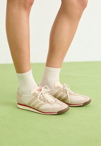 Zapatillas deportivas beige con parte superior de malla, tres franjas laterales, punta redonda y acentos rojos en la suela, combinadas con calcetines de color claro sobre una superficie verde.