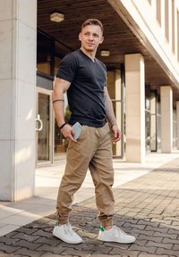 Gray V-neck t-shirt, tan jogger pants with elastic cuffs, and white sneakers with a green accent. Standing outdoors, holding a phone.
