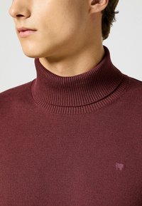 Burgundowy sweter typu golf z materiału prążkowanego. Posiada wysoki kołnierz oraz mały akcent z logo na klatce piersiowej.